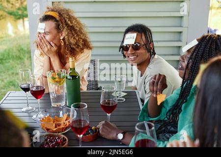 Group of young people laughing and playing Guess who game while sitting at table outdoors in Summer Stock Photo