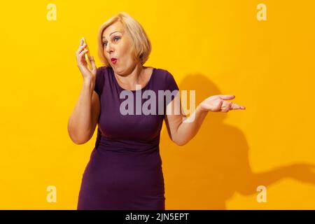 Emotional senior woman in purple dress using cellphone isolated on bright yellow background. Concept of beauty, age, emotions, modern gadgets Stock Photo