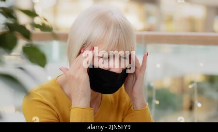 Close-up unhappy caucasian middle-aged woman in medical mask sitting indoors holding hands behind head suffering headache closing eyes feeling tired Stock Photo