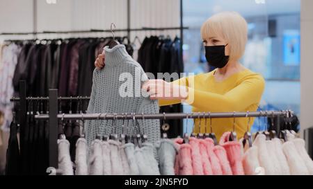 Mature focused woman shopper in protective mask searching clothes in boutique attractive middle-aged lady customer in clothing store looking for new Stock Photo