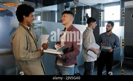 Young diverse people students stand in corridor near classroom communicate talking discussing topic of test prepare read notes taking exam waiting for Stock Photo