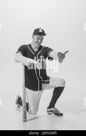 Portrait of young cheerful man, baseball player standing on one knee, leaning on bat and posing. Black and white photography Stock Photo