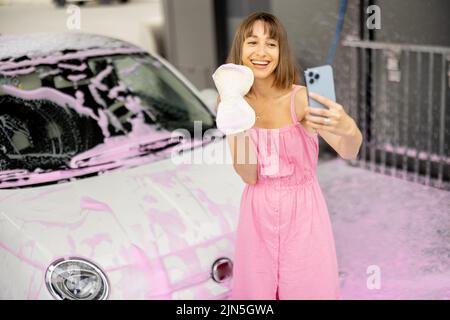 Woman in pink dress takes selfie on phone while washing her car Stock Photo