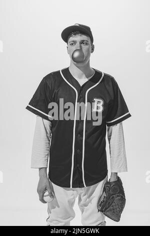 Portrait of young man, baseball player in uniform with bubblegum, in glove with ball posing. Black and white photography Stock Photo