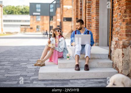 Stylish colleagues work on laptop and eat take away food on a street outdoors Stock Photo
