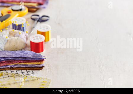 Ruler, cutting mat, stack of multi-colored pieces of fabric, pincushion, spools of thread, scissors, rotary cutter, stack of fabric on a white surface Stock Photo