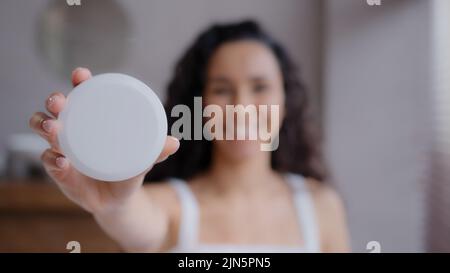 Out focus happy smiling young woman standing in bathroom holding cosmetic product in hand demonstrating jar of skin care cream advertise promotes Stock Photo
