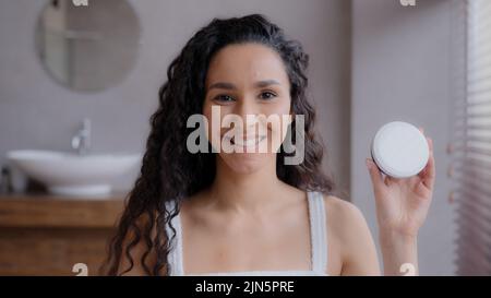 Close-up young attractive happy hispanic woman standing in bathroom looking at camera smiling advertising skin care product pointing at jar Stock Photo