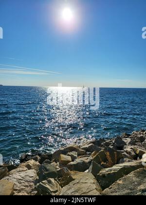 sun is high over the sea with stony coast and light reflections on the water surface Stock Photo