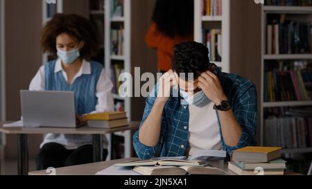Tired overworked young arab male student in protective mask sits at desk in library does homework writes notes prepares for exam makes mistake grabs Stock Photo