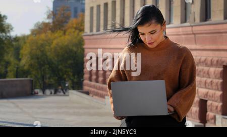 Young businesswoman freelancer student sitting outdoors reading email on laptop smiling laughing good news feels happy enjoys process surf web on Stock Photo