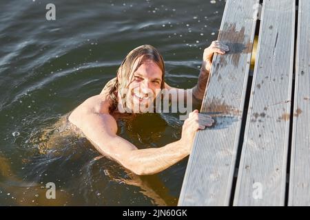Portrait of long haired young man smiling at camera while swimming in lake lit by sunlight Stock Photo