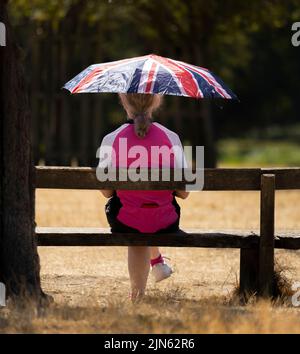 Woman sitting on park bench with umbrella sheltering from strong sun in a heatwave, London, UK Stock Photo