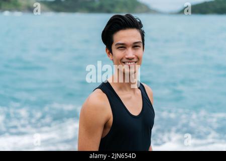 Young happy smiling Eurasian man nearby the sea and looking at the camera. 20s healthy Asian guy in tank top with relaxation. Stock Photo