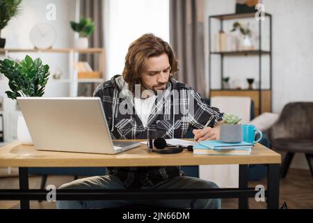 Focused bearded male student in casual wear sitting at table with modern laptop writing notes. Concept of people, technology and distance learning. Stock Photo