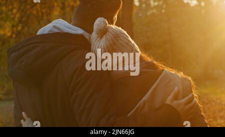 View from back close-up young couple in love in autumn park hugging cute chatting talking enjoying pleasant pastime together admiring view of sunset Stock Photo