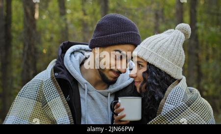 Young happy married couple on date in nature guy and girl lovers in autumn forest embracing in warm blanket drinking tea communicate talk cute hugging Stock Photo