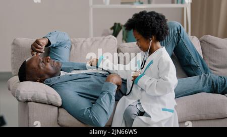 Little cute kid girl plays pretending be doctor adult father lies on couch complains pain symptom playful daughter listening dad with stethoscope Stock Photo