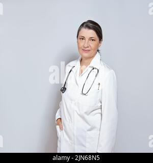 Middle aged female doctor in a white lab coat, smiling friendly, on neutral gray or grey background, copy or text space. Stock Photo