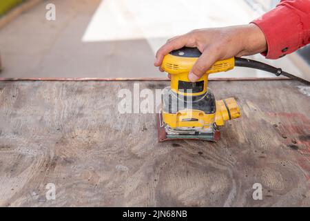 Close-up of a man's hand sanding a piece of wood with an electric sander on a patio outdoors Stock Photo