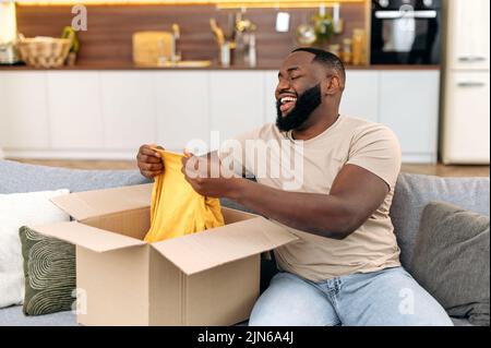 Excited Happy smiling african american guy unpacks his long awaited package, examines the clothes, happy with a good purchase, sits at home on the sofa in the living room. Online shopping concept Stock Photo