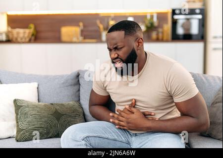 Unhappy guy of african american ethnicity, sits on a sofa in the living room, holds his hands on his stomach, grimaces from pain in his stomach, suffers from poisoning, spasm, stomach problems Stock Photo