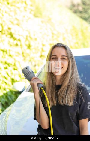 Vertical view of young beautiful woman smiling holding electric car plug in green background as renewable energy concept Stock Photo