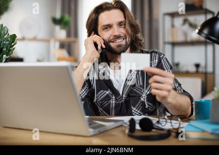 Young bearded man with casual outfit sitting at desk with wireless laptop and showing empty white card. Space for text on business card. Stock Photo