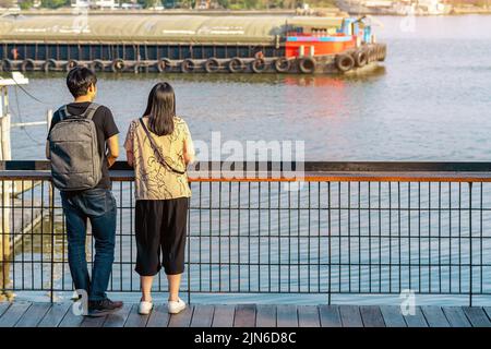 Back view of Asian couple relaxing and chilling at a riverside. Relaxed lifestyle for lover enjoying freedom in summer holidays. Happy people outdoors Stock Photo