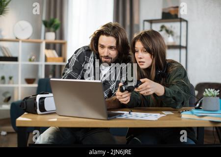 Young happy couple playing video games at home, sitting at the table with laptop and using joysticks. Happy man in headset shows new game for his lovely smiling girlfriend at home. Stock Photo