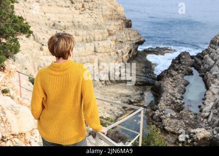 Elderly tourist woman watching waves on rocks during sunset Stock Photo