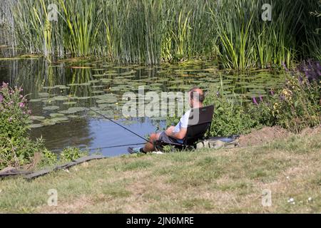 FIsherman with rod and line relaxing by lil;y-covered inlet on River Avon Weston on Avon UK Stock Photo