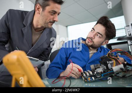 supervisor talking to electronics apprentice using multimeter Stock Photo
