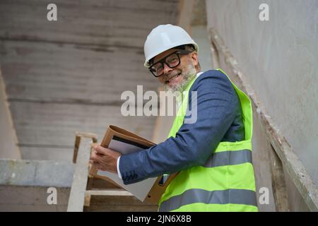 Smiling man foreman holding folder while standing on stairs in cottage Stock Photo