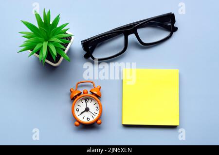 Adhesive Note with potted plant and alarm clock set at 8 o'clock against blue background. Business concept. Copy space. Stock Photo