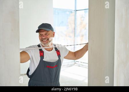 Joyful gray-haired builder measures width of doorway with tape measure Stock Photo