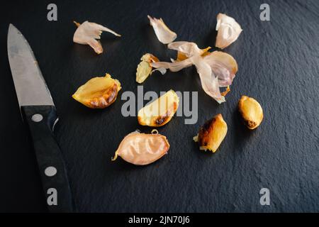 Roasted Garlic Cloves on a Dark Slate Stone Background: Peeled and unpeeled roasted garlic with a paring knife Stock Photo