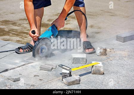 A worker using an electric grinder and a diamond cutting blade cuts paving slabs while laying pavement on a hot afternoon. Copy space. Close-up. Stock Photo