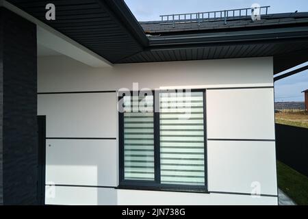 Corner of modern house with shingled roof and big window with closed shutters on white wall Stock Photo