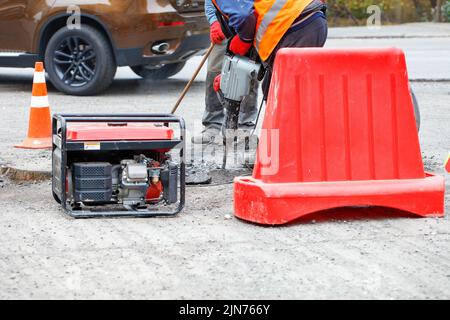 Road workers are repairing the carriageway of a section of the road with an electric jackhammer fenced with a red road shield. Stock Photo