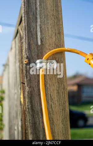 Yellow extension cable through metal bracket screwed to the support pole of a wooden fence. Blurred fence and house in the background Stock Photo