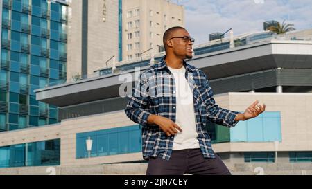 Stylish happy cheerful afro american guy african man singing song sings pretend playing guitar drums musician enjoying music dancing outdoors city Stock Photo