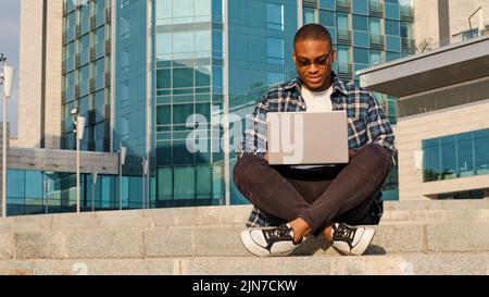 Concentrated young business man african american male freelancer guy student in sunglasses sitting city background company building with laptop Stock Photo