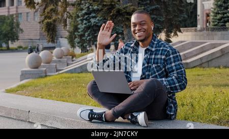 Successful african smiling man guy student male freelancer programmer designer in wireless headphones sitting on terrace in city with laptop working Stock Photo