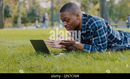 African american guy young business man user lying on green grass lawn in park using laptop online call conference chat remote conversation distant Stock Photo