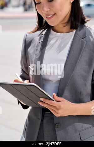 Young smiling Asian entrepreneur standing on city street using digital tablet. Stock Photo