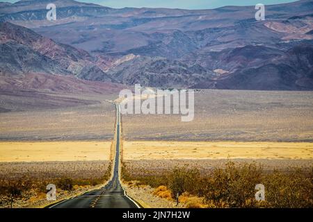 Two lane blacktop highway wavering in the heat  stretching into the distant mountains with band of sand intersecting the rough textured sagebrush terr