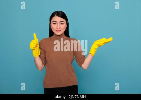 smiling happy cheerful asian young woman from chores pointing hand aside at copy space and wearing yellow glove, Cleaning home concept, Optimistic satisfied positive female thumbs up Stock Photo
