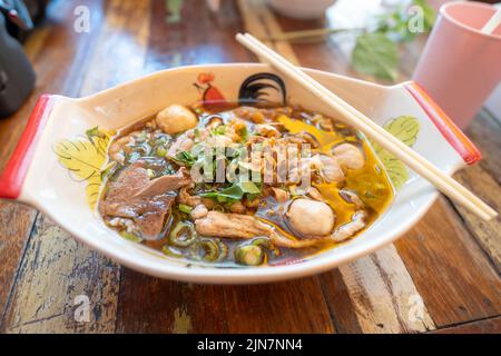 Thai noodle with pork, meatball, pork liver with blood soup in bowl that look like boat on the wood table. Stock Photo