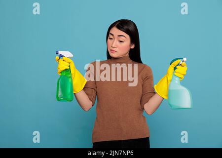 Portrait of pretty sad asian woman holding two detergent sprayers in yellow gloves isolated on a blue background, Cleaning home concept, Unhappy depressed and negative housewife Stock Photo